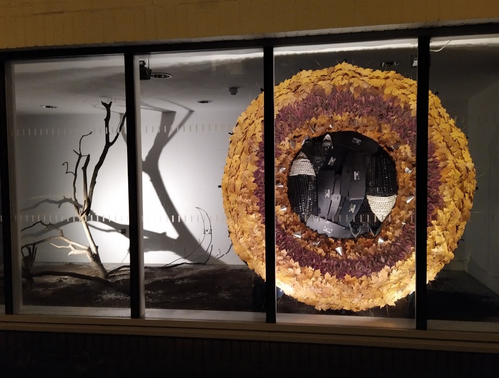 View of the sculpture, which features a concave structure covered in leaves, with two foxes made out of keyboards nestled together inside. A starkly lit branch (to look like a winter tree) is to the left. 